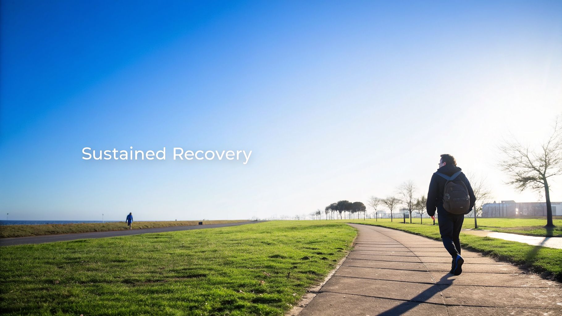 A person with a backpack walks on a sunny path, with green grass and a wide blue sky, conveying sustained recovery.